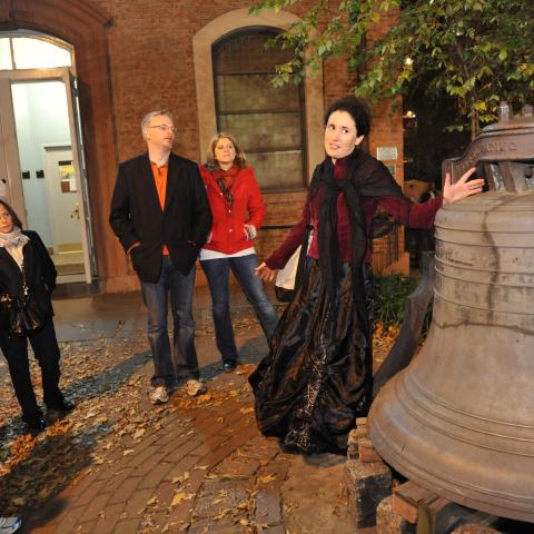 Tour guide in costume speaking to a group near a large outdoor bell at night.