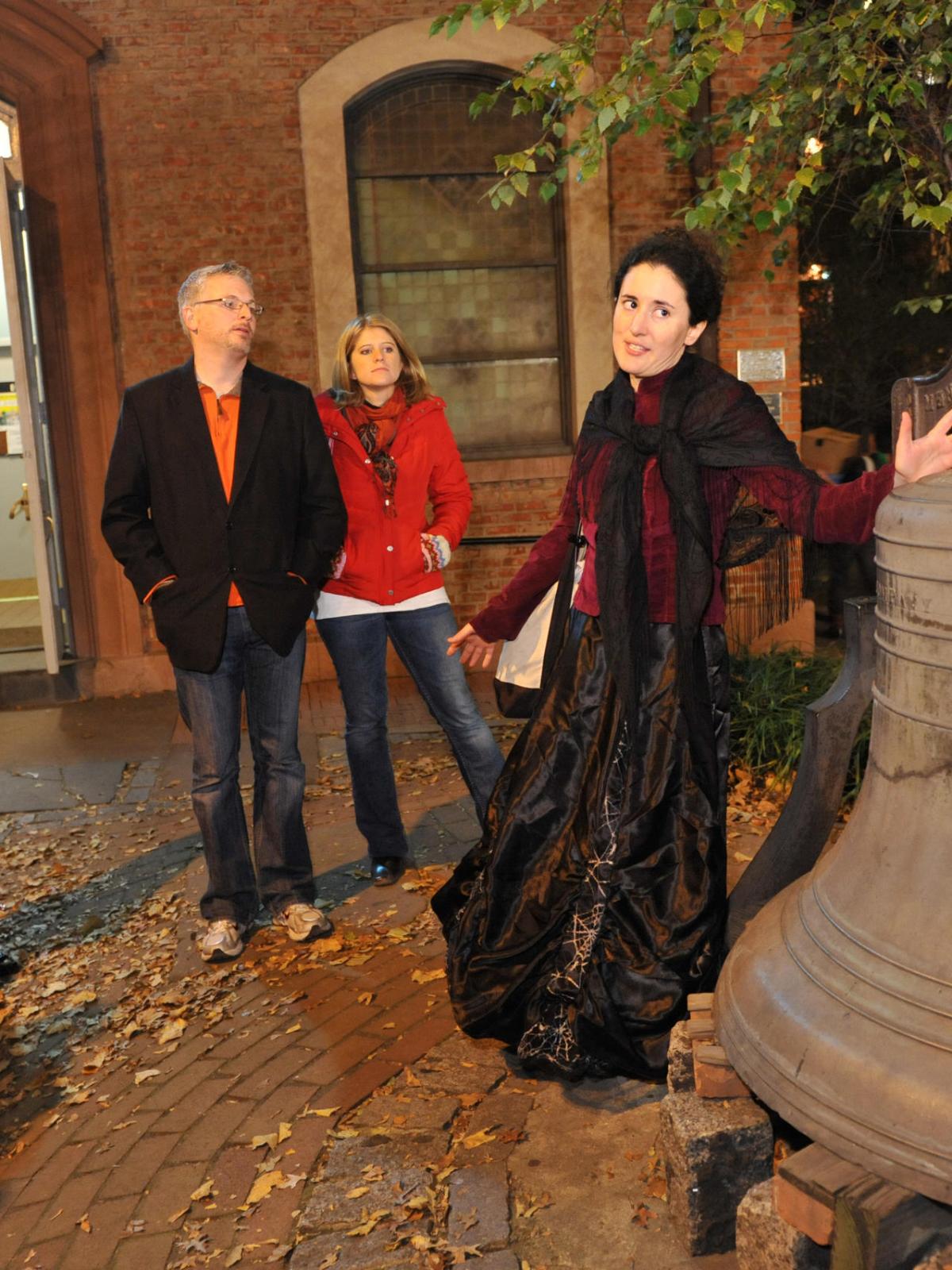Tour guide in costume speaking to a group near a large outdoor bell at night.