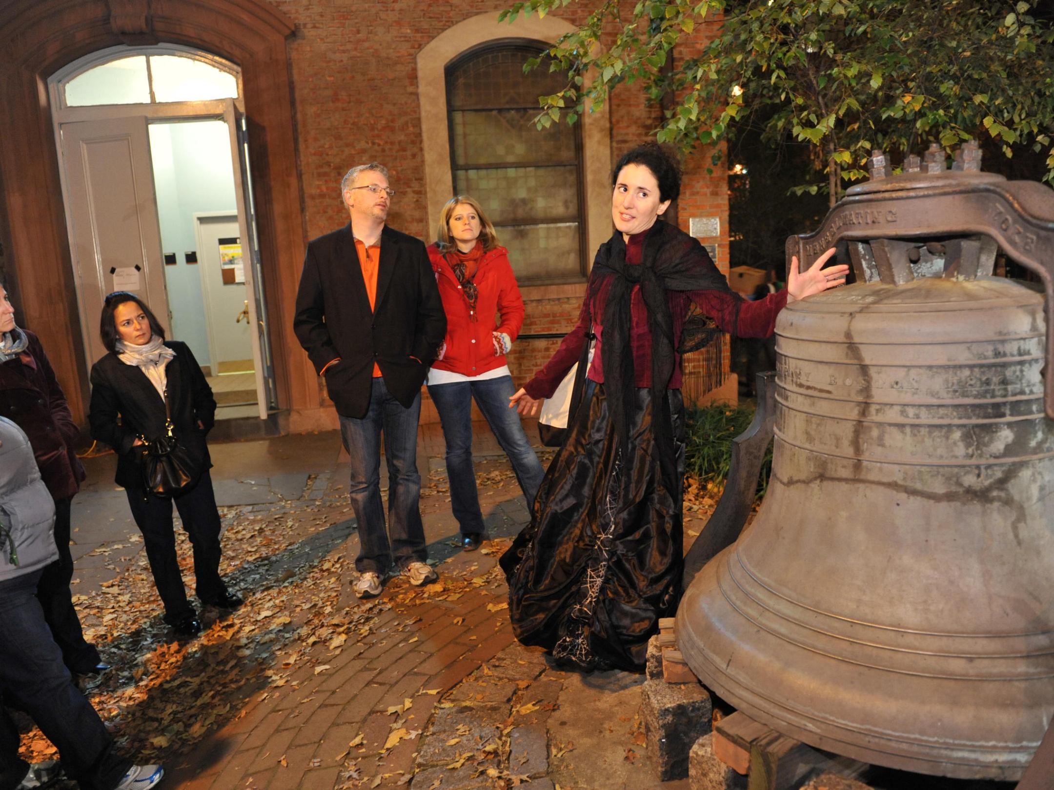 Tour guide in costume speaks near a large bell to a group outside a brick building at night.