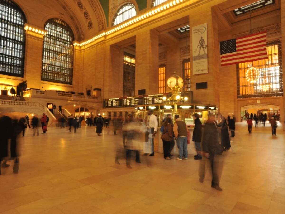 Busy station interior with large clock, American flag, and people walking.