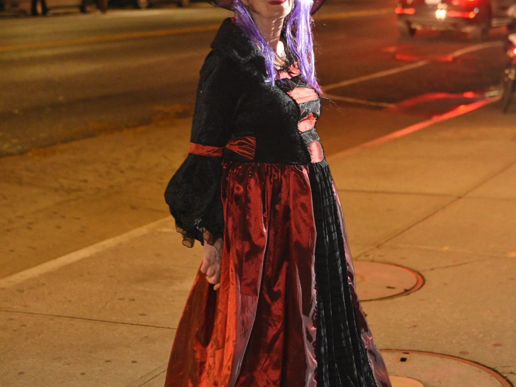Person dressed as a witch with a black hat and gown, standing on a city street at night.