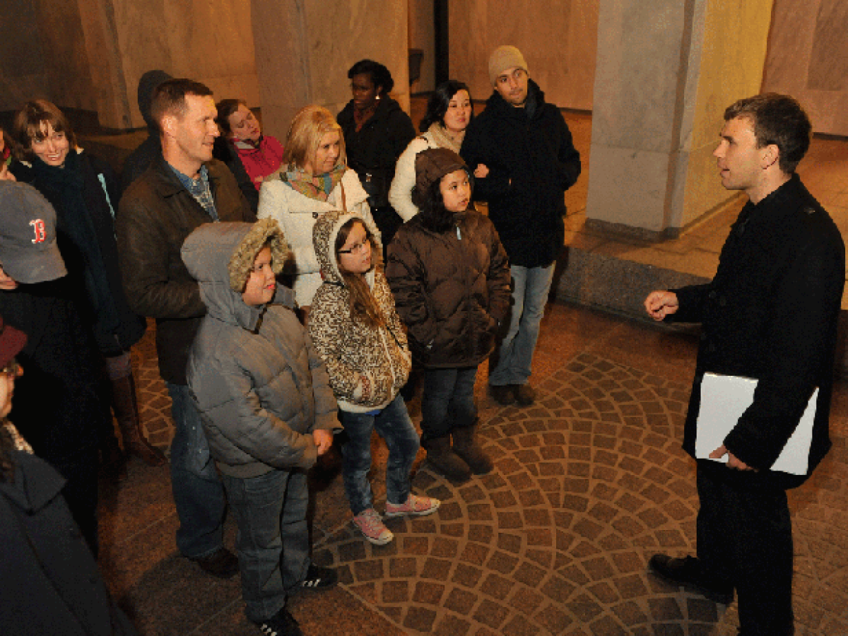 A man speaks to a group of people, including children, in a dimly lit indoor area.