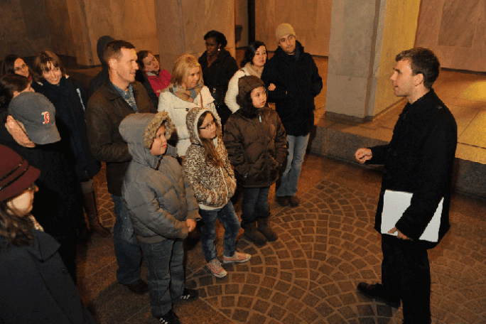 A man speaks to a group of people, including children, in a dimly lit indoor area.