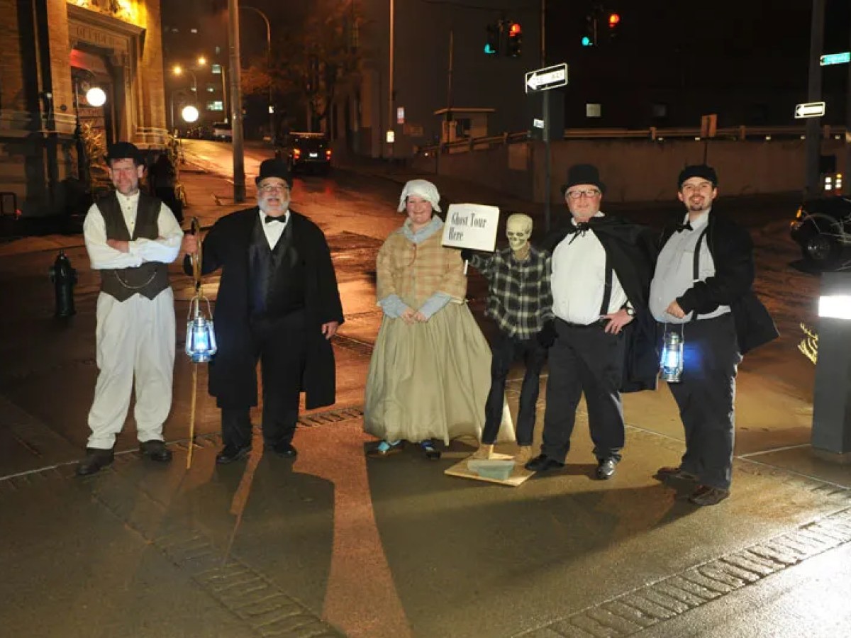 Group in historical costumes with lanterns, next to a skeleton with a 'Ghost Tour Here' sign, on a city street.