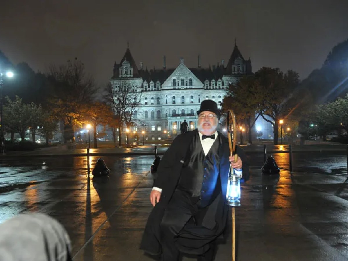 Man in vintage costume with lantern in front of illuminated historic building at night.