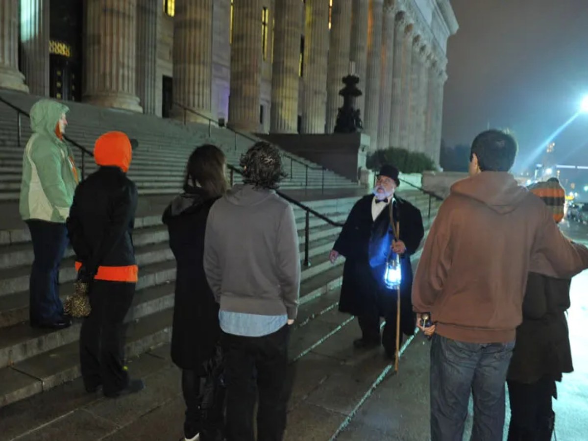 Group of people on a night tour by a historic building, led by a guide with a lantern.