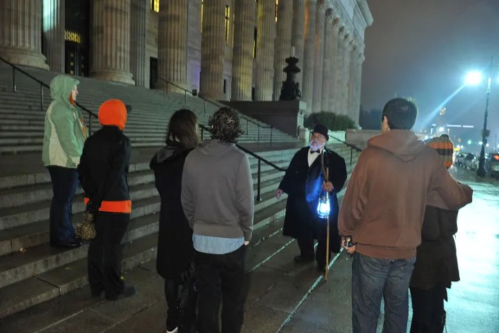 Group of people on a night tour by a historic building, led by a guide with a lantern.