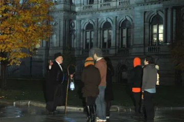 People gathered at night near a historic building with lit windows and autumn trees.