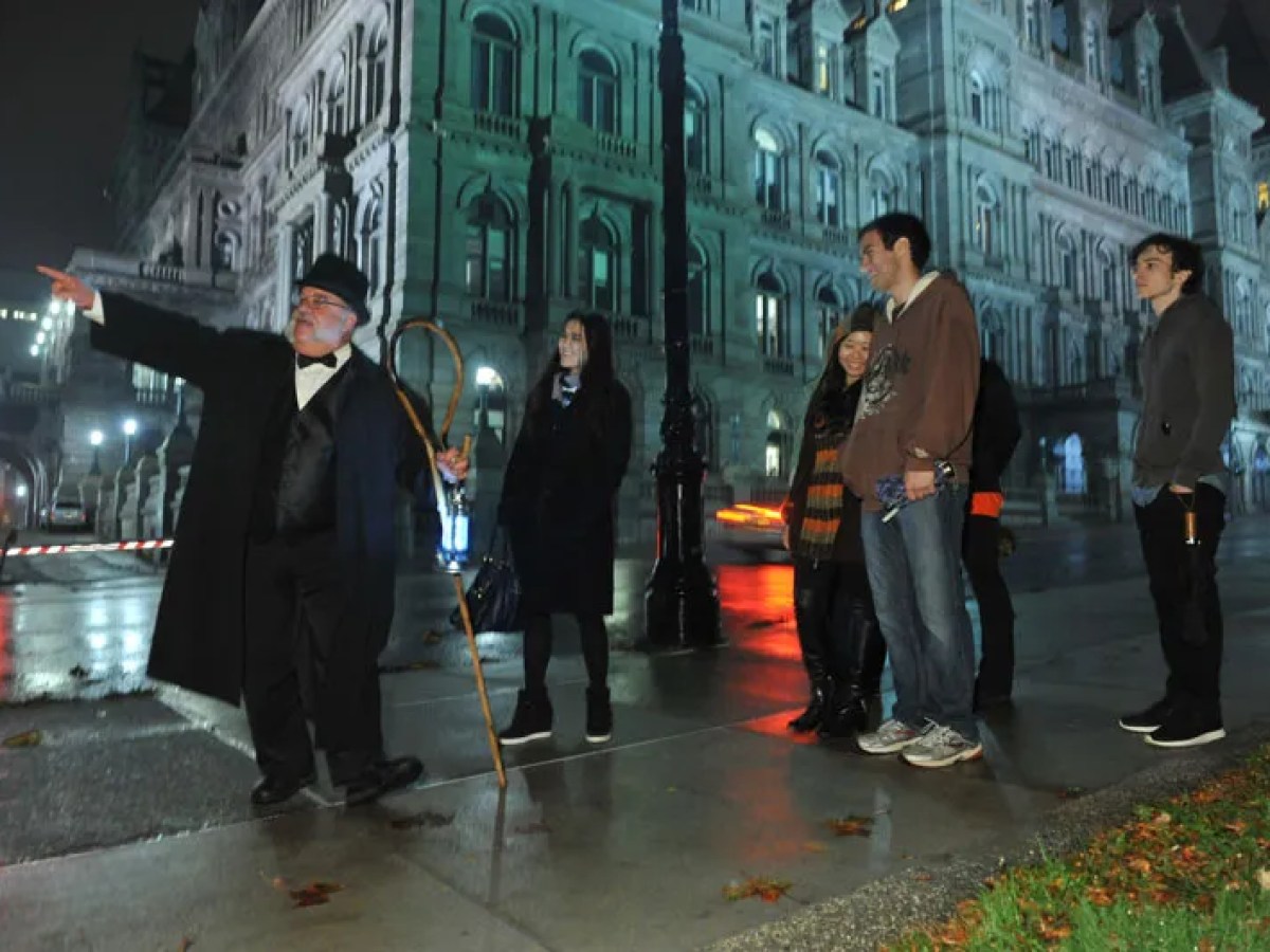 Tour guide points out a building to a group of people at night on a rainy street.