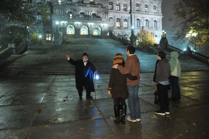 Tour guide with lantern speaks to group near illuminated grand staircase at night.