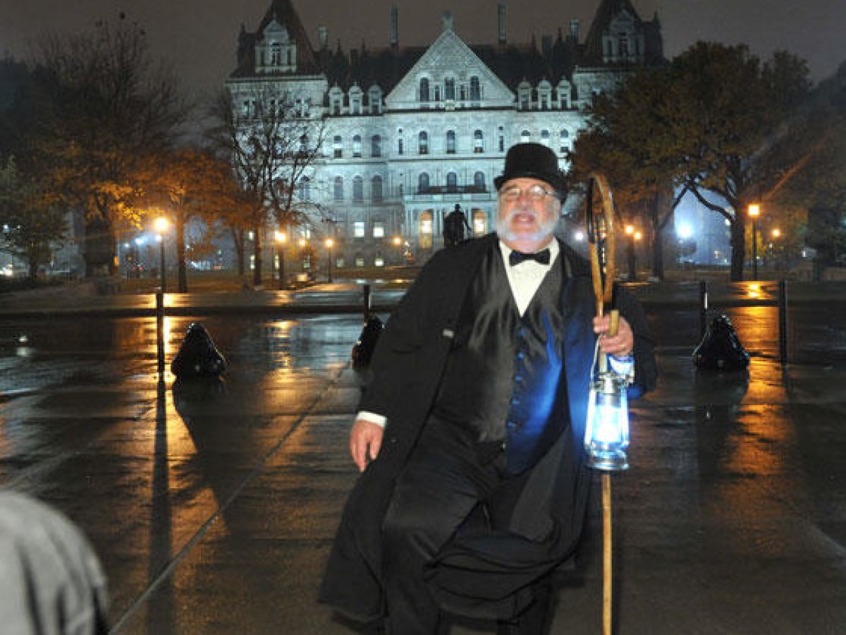 Man in formal attire and top hat holds lantern, standing on wet pavement with illuminated building behind.