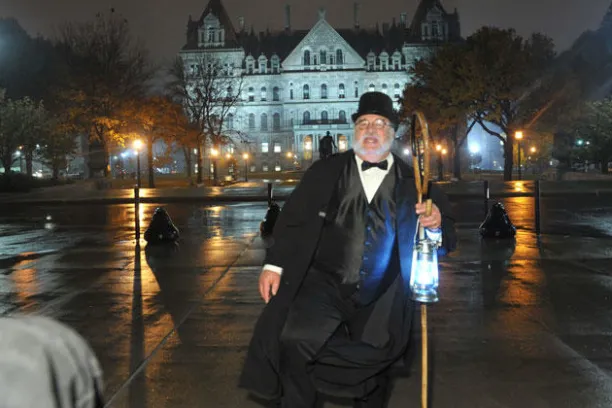 Man in formal attire and top hat holds lantern, standing on wet pavement with illuminated building behind.