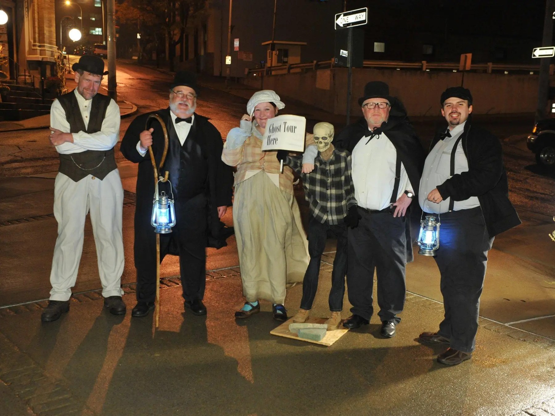 Group in costumes with lanterns on a night street, one holding 'ghost tour here' sign.