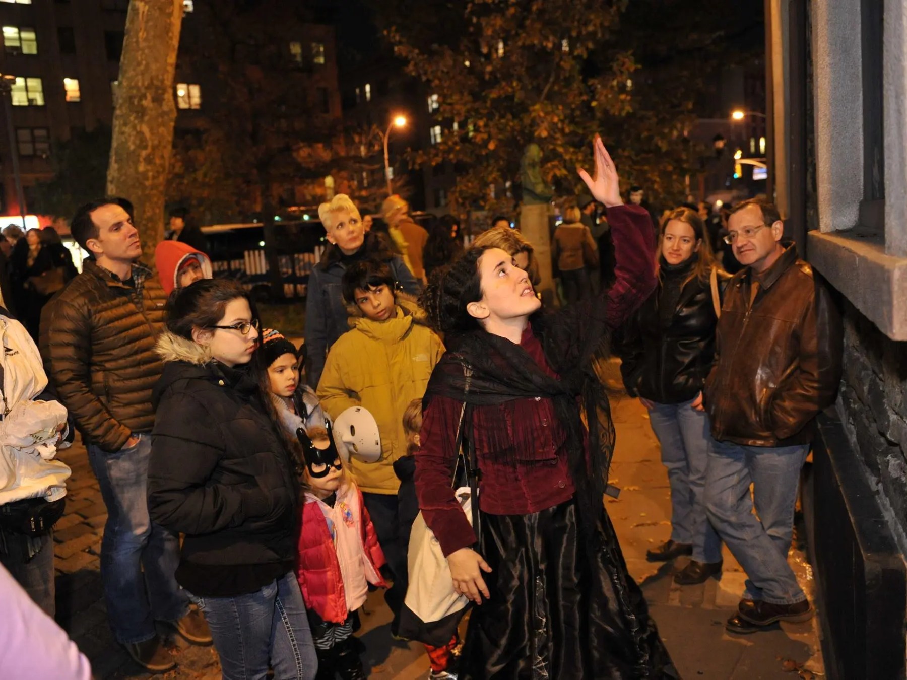 Woman in period costume gestures upward, surrounded by a nighttime tour group.