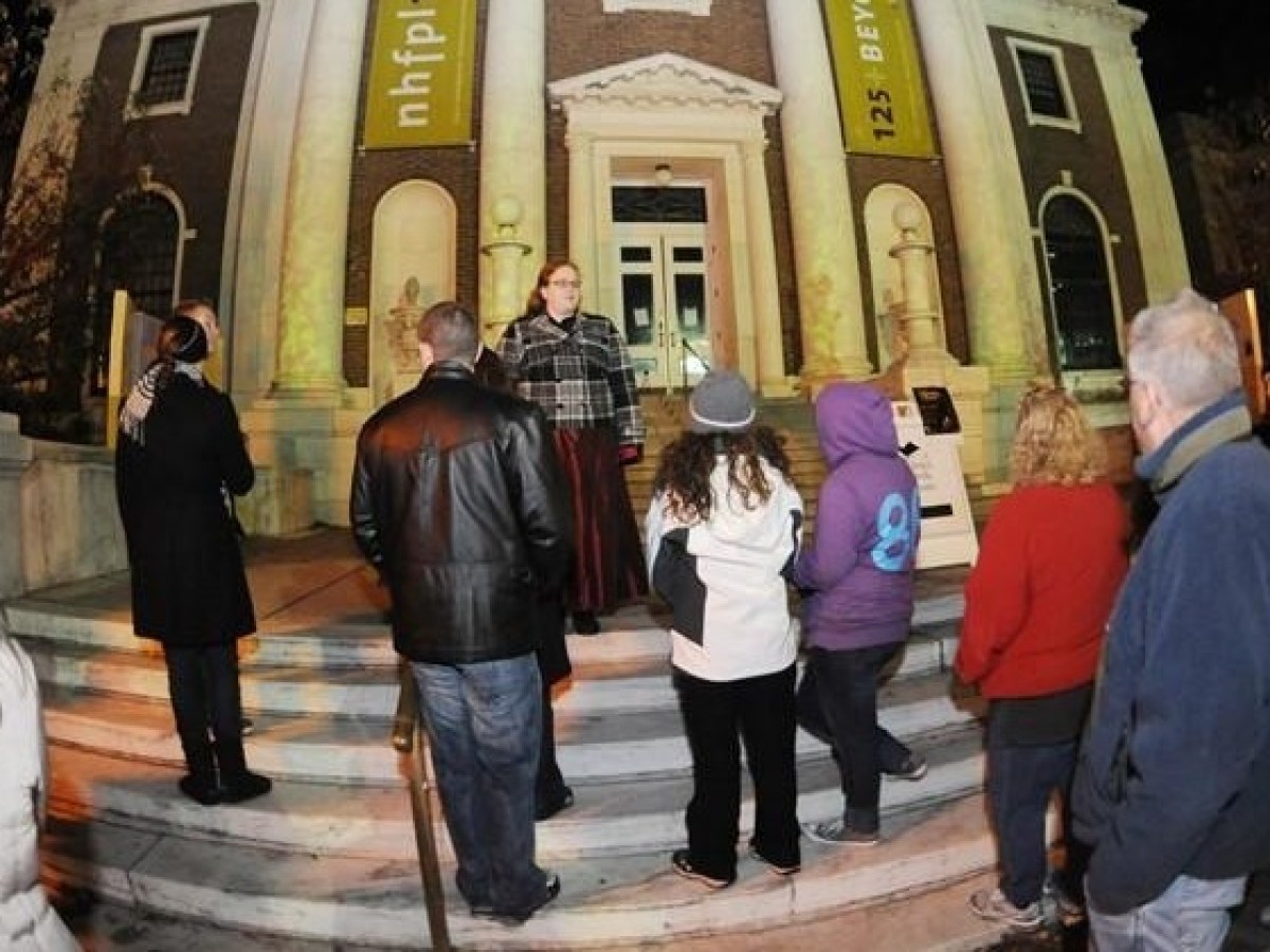 Group of people gathered outside a historic building at night, listening to a person speak on the steps.