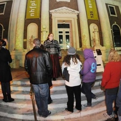 Group of people gathered outside a historic building at night, listening to a person speak on the steps.