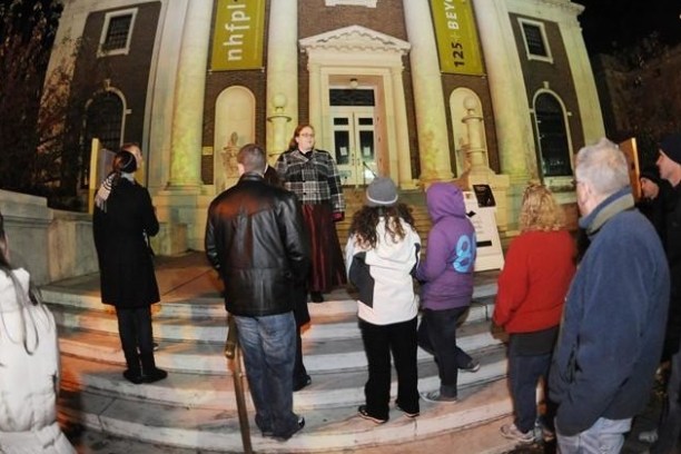 Group of people gathered outside a historic building at night, listening to a person speak on the steps.