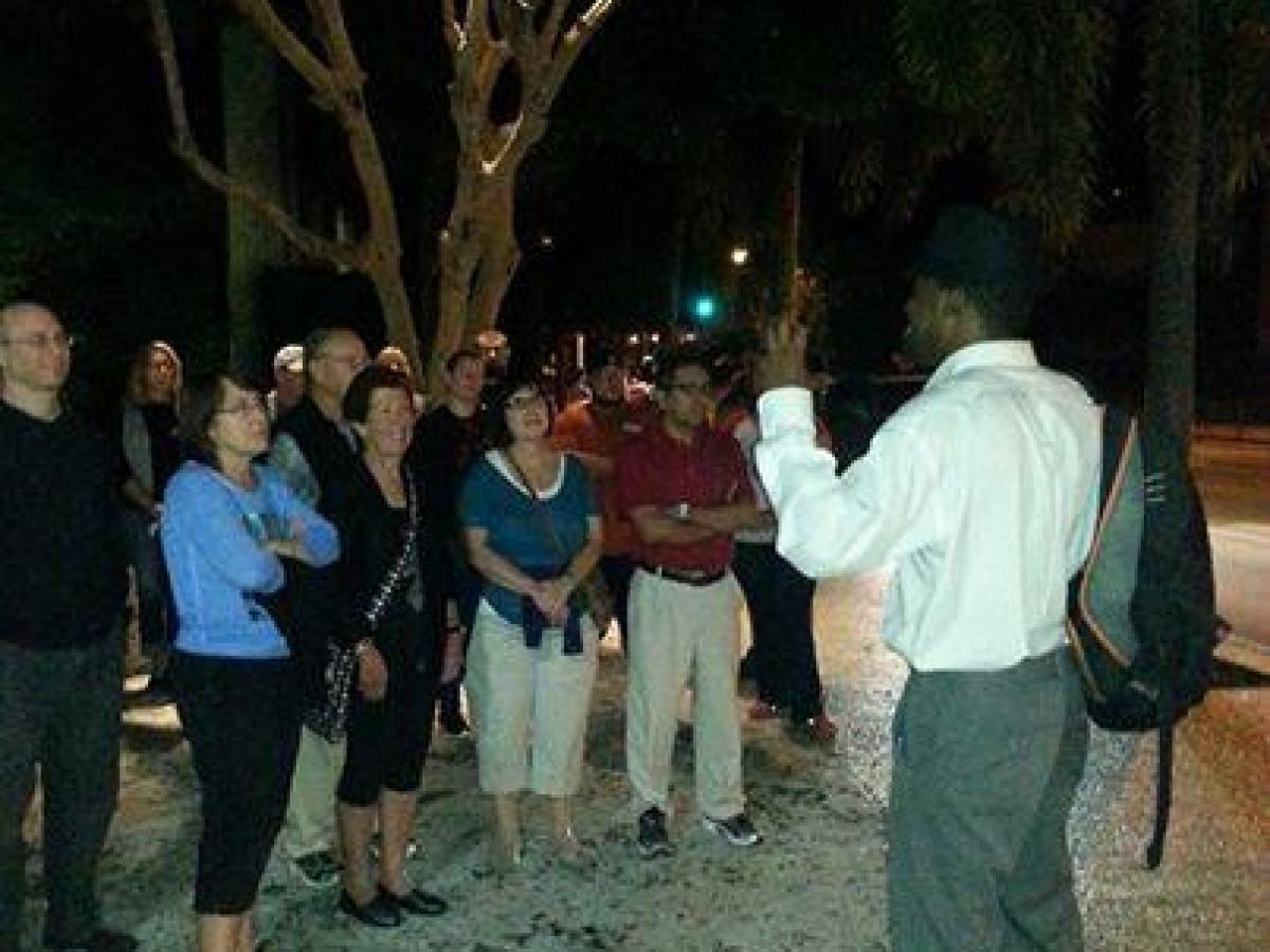 Person speaking to a group outdoors at night with trees in the background.