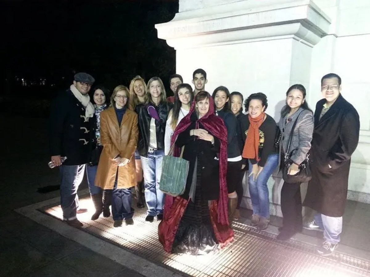 Group of people standing together at night near a lit area on the ground, one in a red cloak.