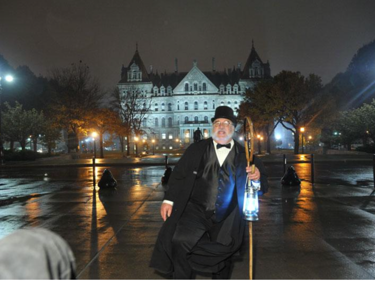 Man in old-fashioned attire holding lantern, standing on wet pavement at night with illuminated building in background.