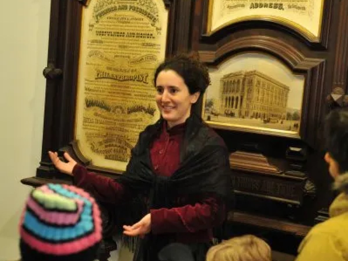 Person in period costume speaking to group in front of historical plaques.
