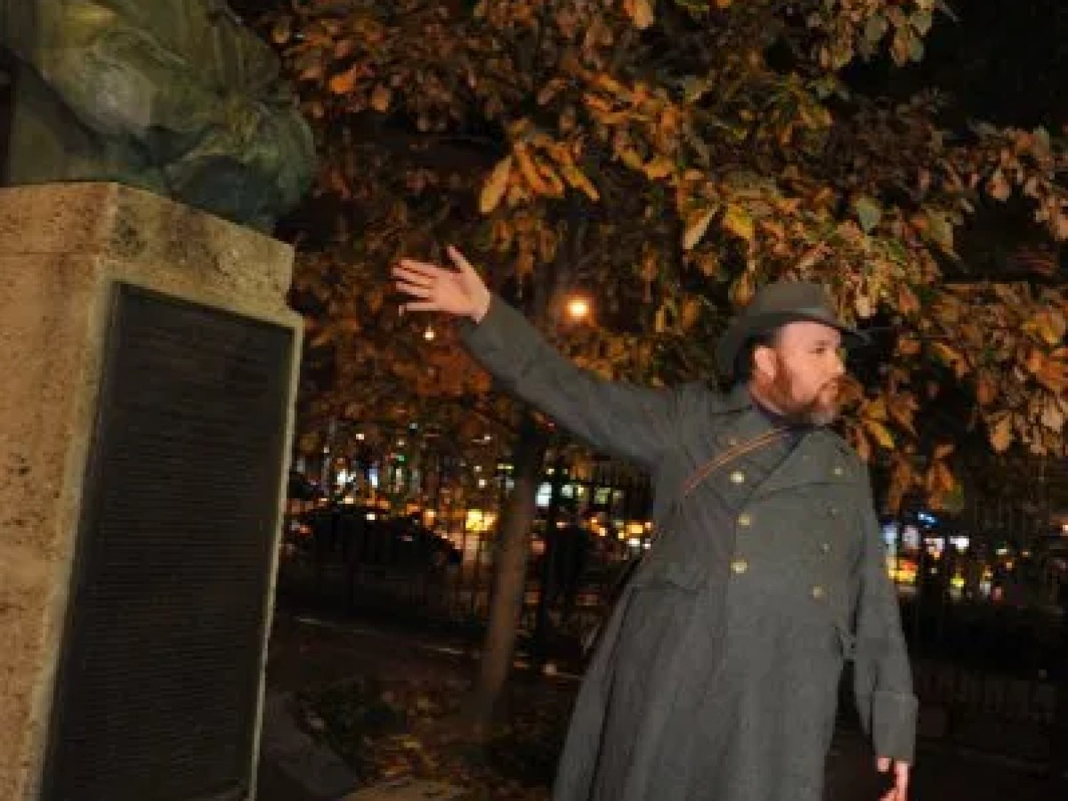Person in a long coat gestures next to a statue at night, with trees and city lights in the background.